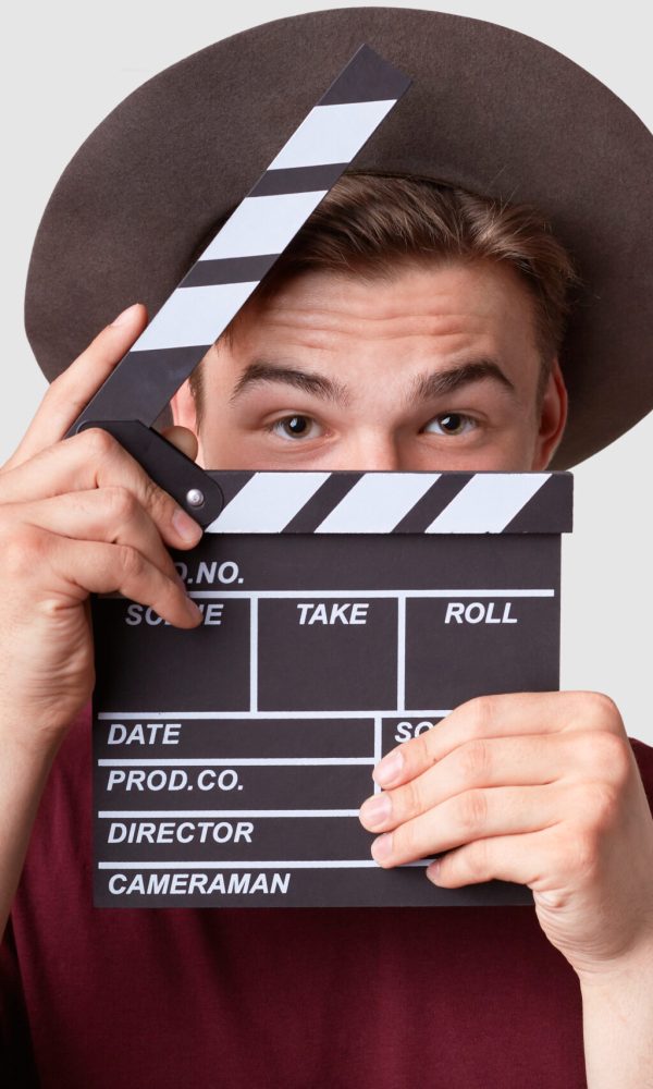 Caucasian successful male producer looks through oprnrd clapperboard, works on film production, wears hat and casual t shirt, isolated over white background. Movie industry and cinematography concept